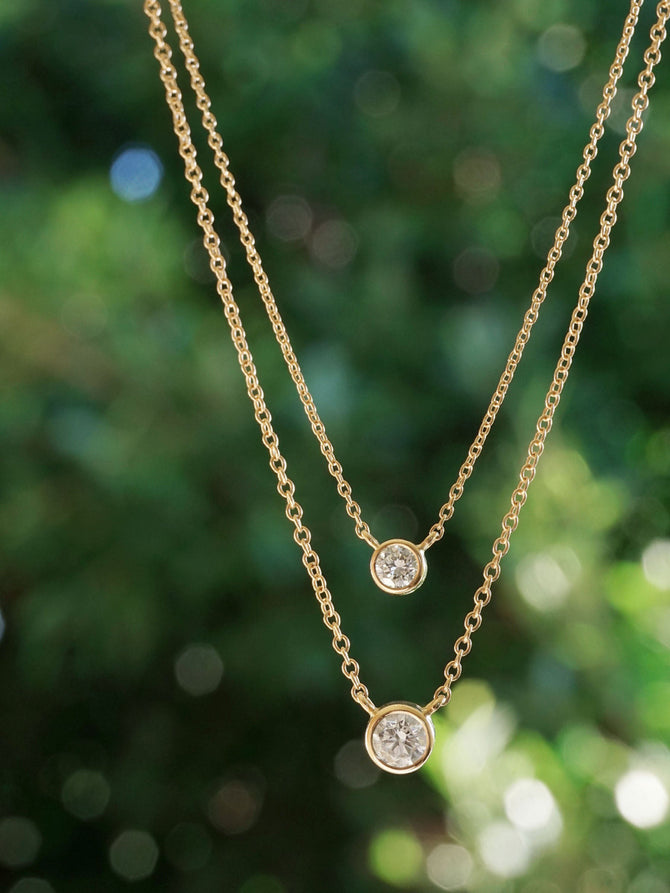 Two gold necklaces with 1/4ct and 1/8ct diamond pendants against a blurred green background