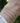 Three delicate bracelets worn on a wrist: a thin gold chain, a gold paperclip chain, and a silver paperclip chain, shown against a blurred green outdoor background.