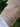 Three delicate bracelets worn on a wrist: a thin gold chain, a gold paperclip chain, and a silver paperclip chain, shown against a blurred green outdoor background.