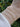 Three delicate chain bracelets on a person's wrist: a fine gold chain, a silver paperclip chain, and a gold paperclip chain, with a white textured sleeve and blurred green foliage in the background.